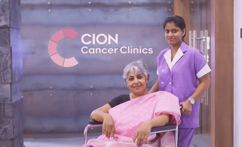 A CION nurse in a lavender uniform beside a smiling patient in a wheelchair, in front of the CION Cancer Clinics logo